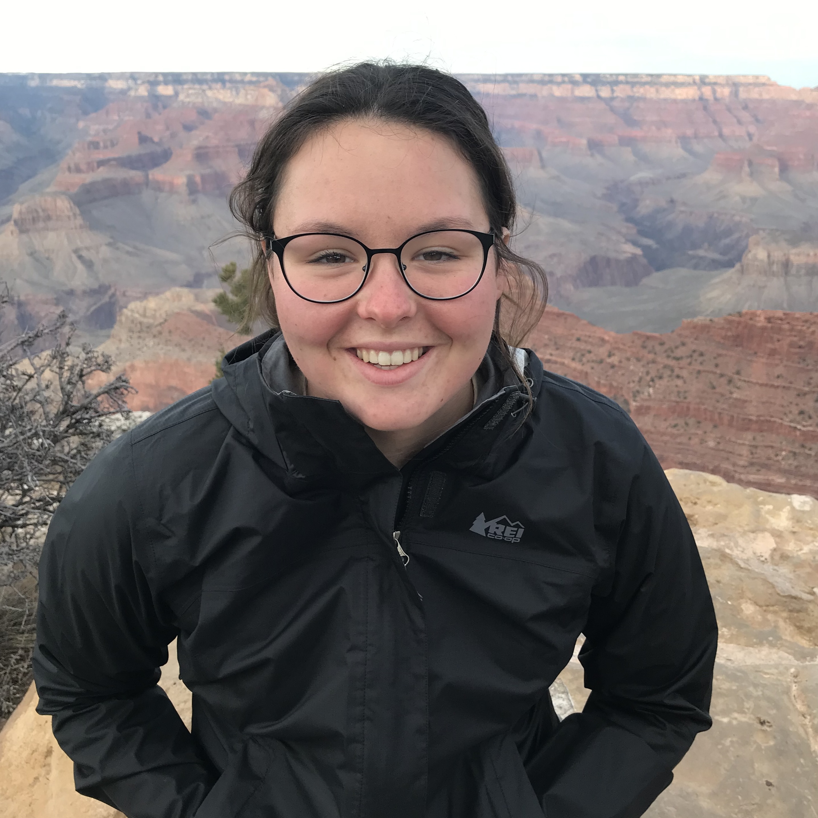 Katie Blessington is a white woman with long dark hair and dark glasses, here she is wearing a parka with the spetacular Grand Canyon as backdrop.