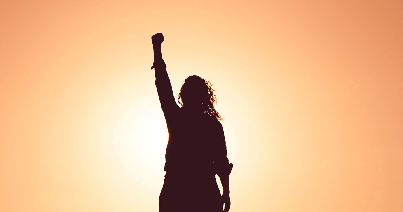 Woman in silhouette with raised fist, facing a rising sun. 